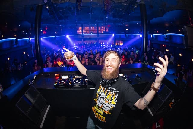 HOTFIRE with both arms raised behind the decks, blue-lit club and full floor behind him